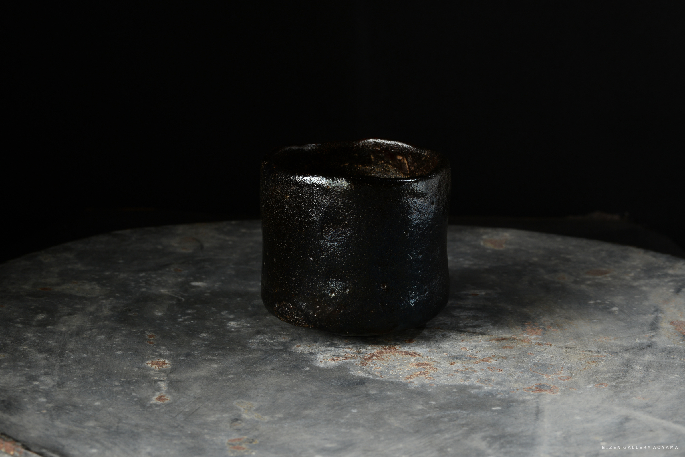 黒い土で作られた陶器のカップが、質感のある石の上に置かれています。背景は暗く、カップに焦点が当てられています。