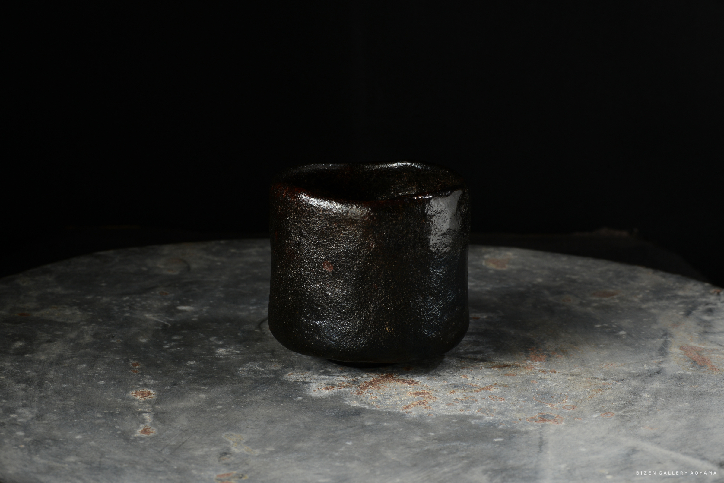 黒い陶器の茶碗が、金属の台の上に置かれています。背景は黒で、茶碗の質感が際立っています。