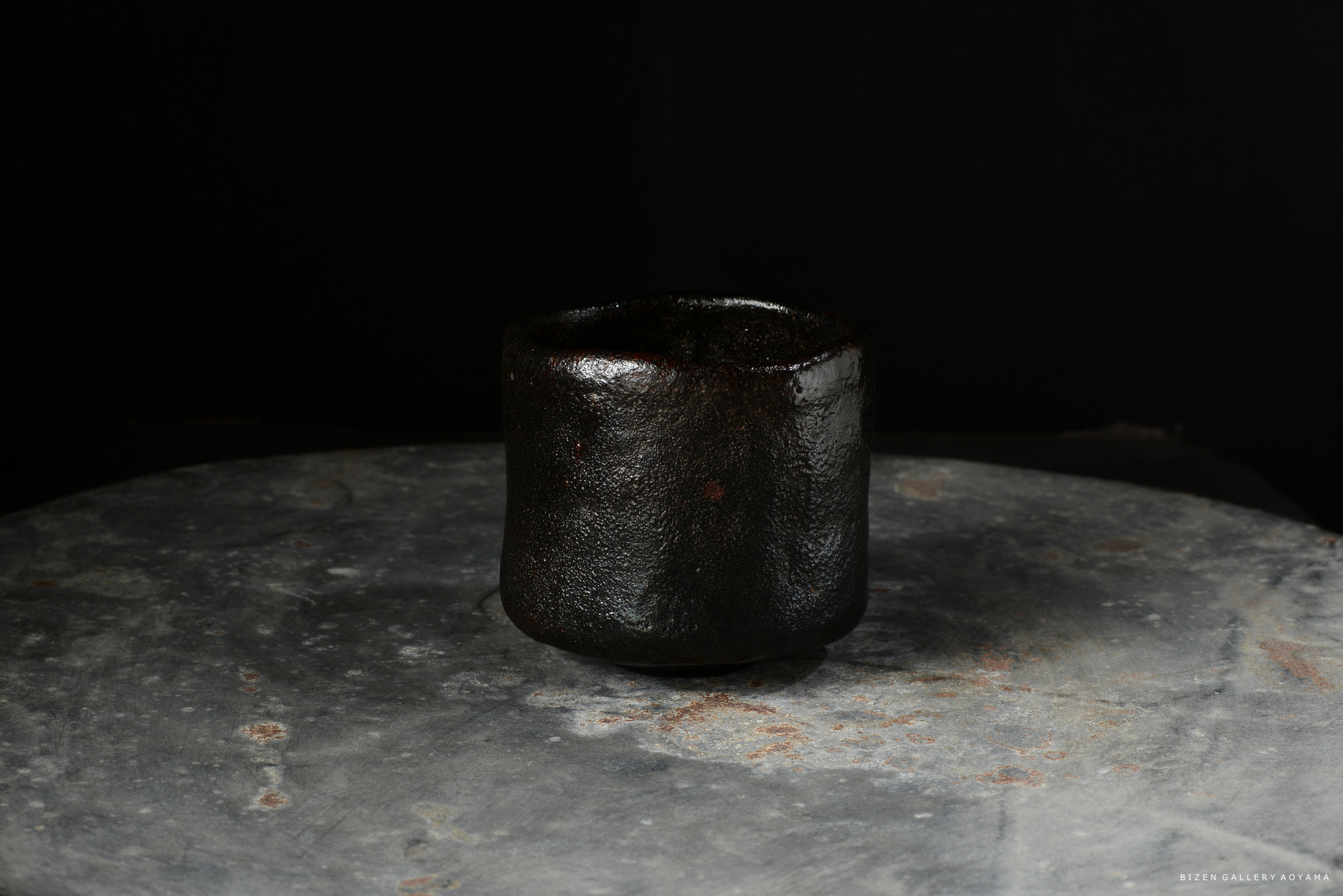 黒い陶器の茶碗が灰色の台の上に置かれている。背景は黒い。