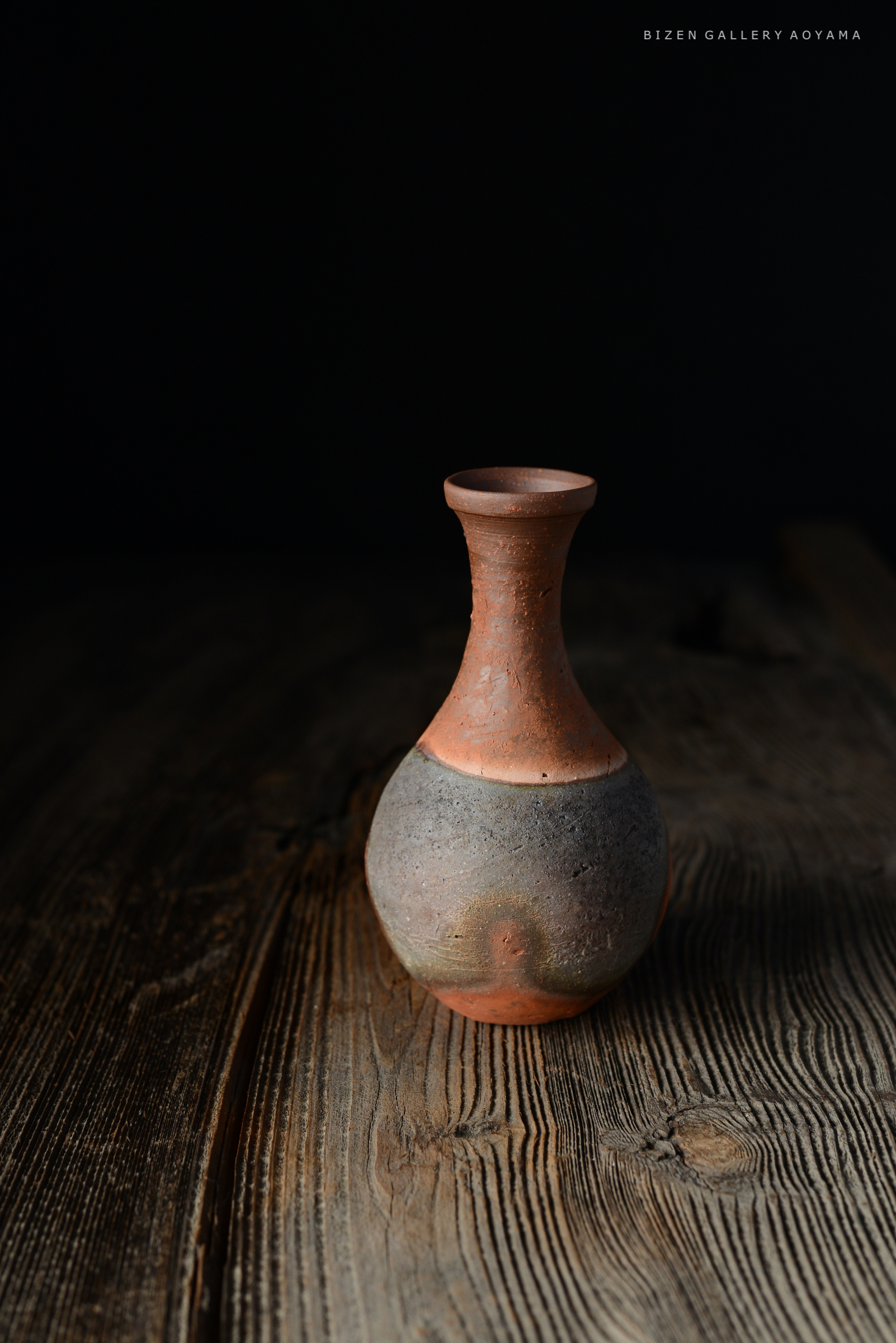 黒い背景の前に置かれた陶器の壺。上部は赤茶色で、下部は灰色の質感を持つ。木のテーブルの上に置かれている。