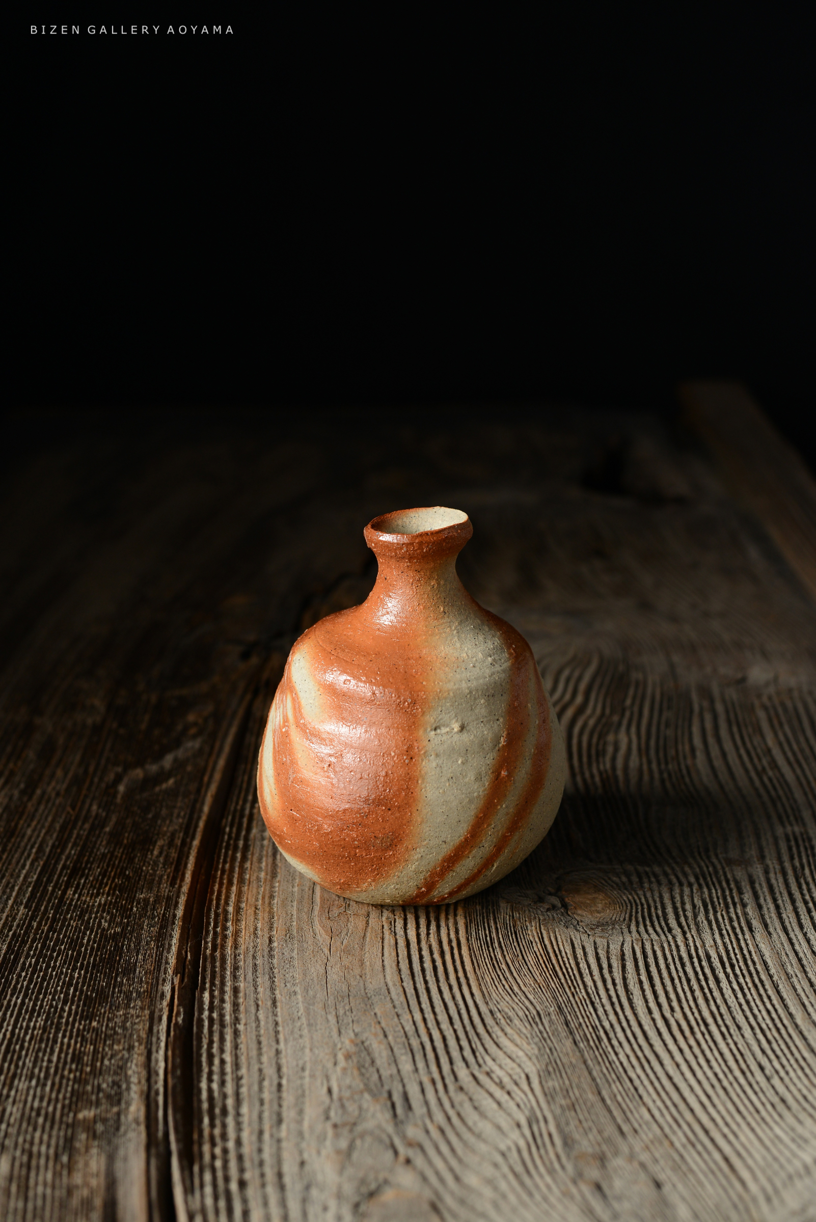 黒い背景の上に置かれた、赤茶色の模様が施された陶器の壺。木のテーブルの上にあり、曲線的な形状が特徴。