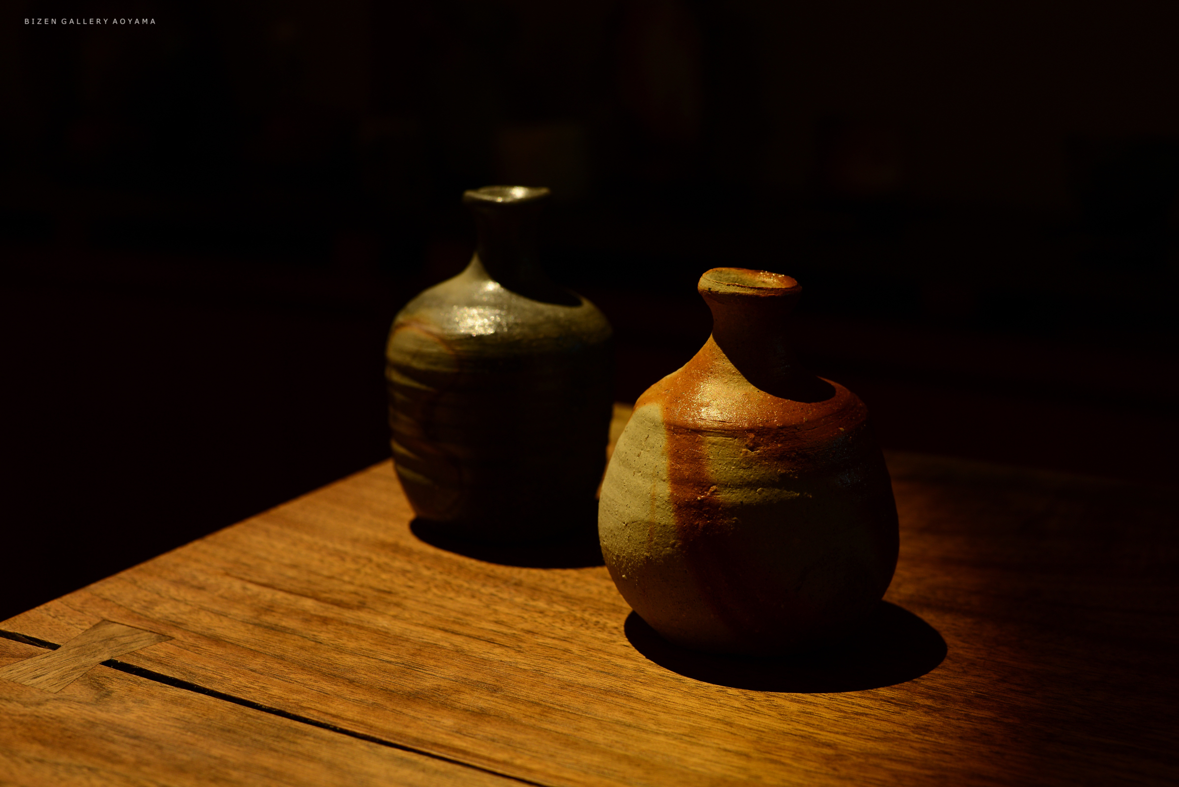 木のテーブルの上に置かれた二つの陶器の壺。左側は黒っぽい色合いで、右側は赤土と少しの灰色が混ざり合ったデザイン。暗い背景の中で、壺が柔らかい光に照らされている。
