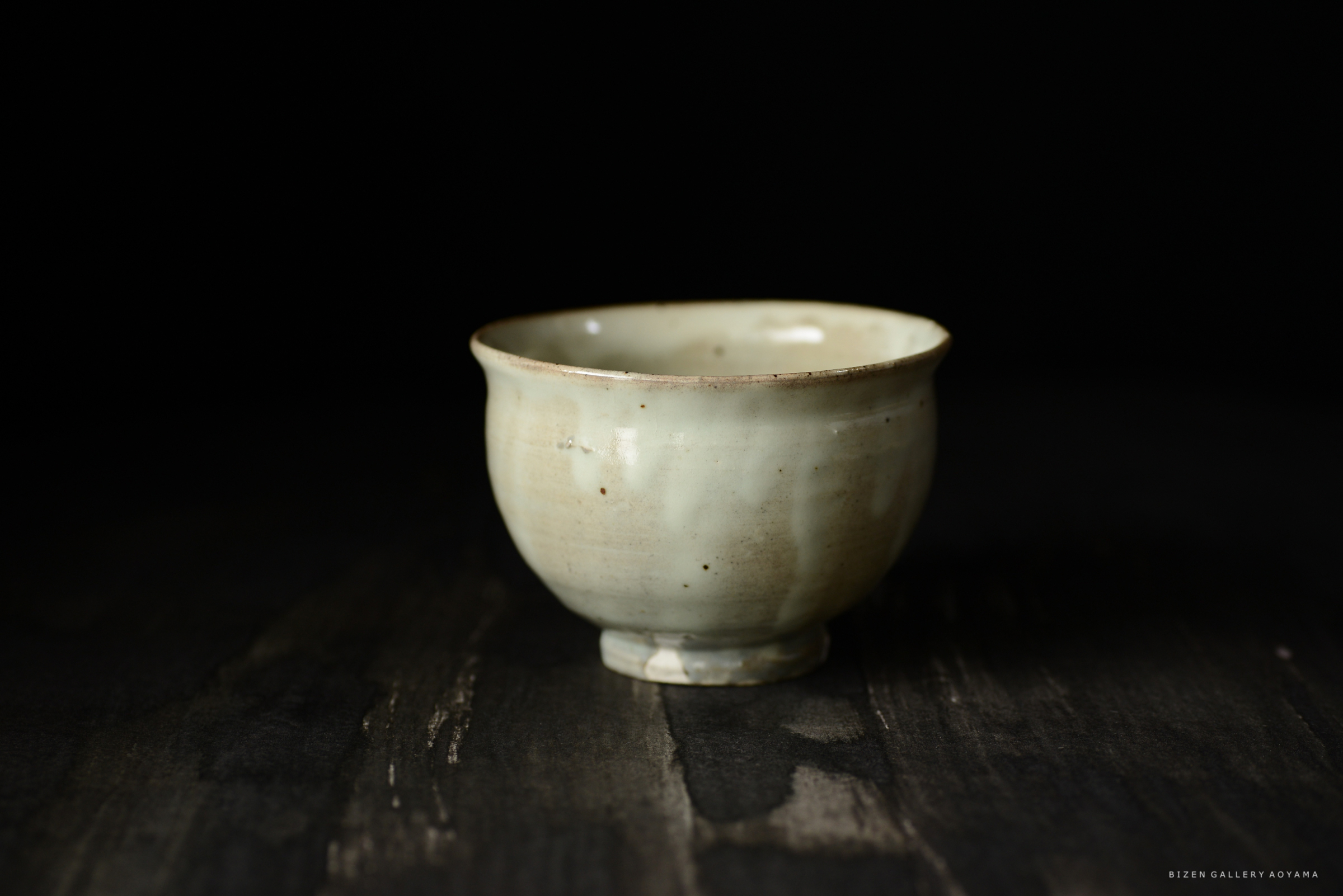 背景が黒色の上に置かれた陶器の小さな碗。淡い緑色で、口縁部分にやや欠けが見える。