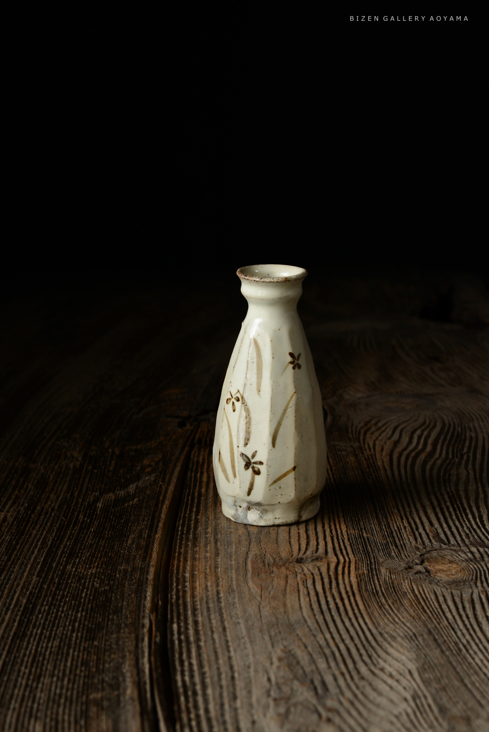 木のテーブルの上に置かれた白い陶器の小瓶。花模様が施されている。背景は黒い。