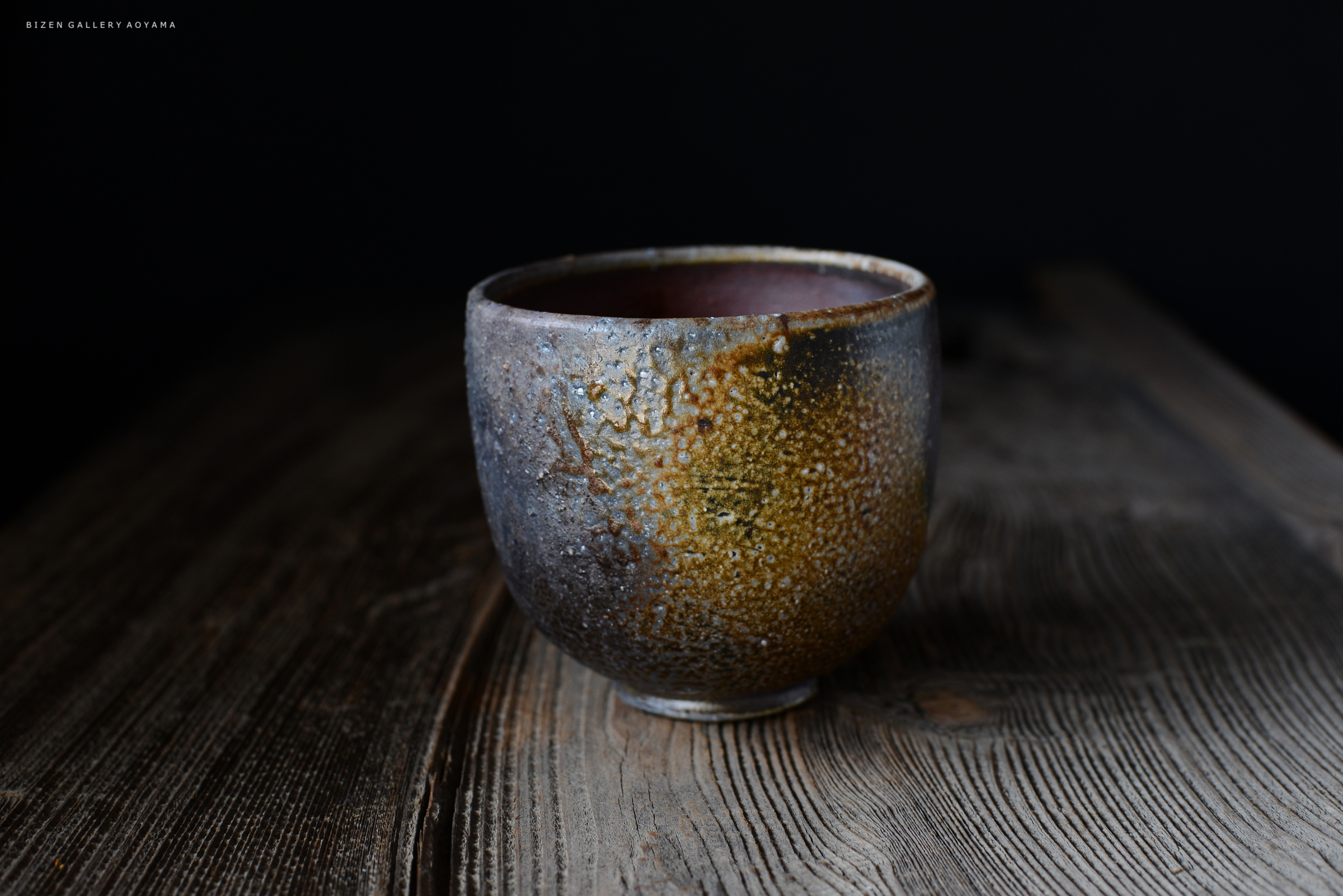 木のテーブルの上に置かれた、質感のある陶器の茶碗。茶碗は、グレーとブラウンの色合いが混ざり合った独特な表面を持っている。
