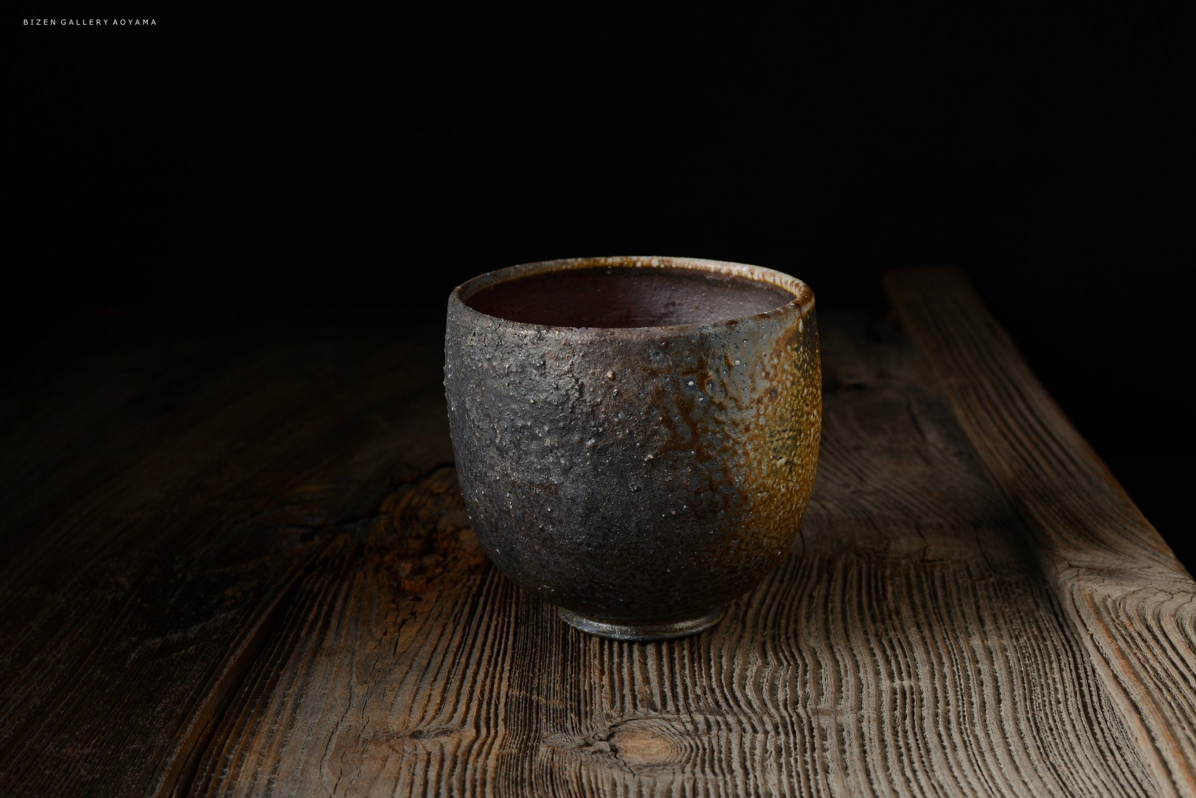 黒い背景の前に置かれた、質感のある陶器の茶碗。木のテーブルの上にあり、独特な釉薬の色合いが特徴。